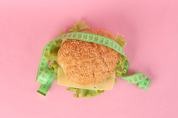 Éhező hamburger mérőszalaggal, rózsaszín háttérrel. Fogyás fogalma - Fotó, kép
