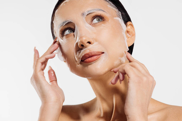 Young beautiful woman in a moisturizing facial mask, skin care. Woman with clean skin using a tissue mask. Woman applying a cosmetic tissue mask to her face on a white background - Foto, Imagem