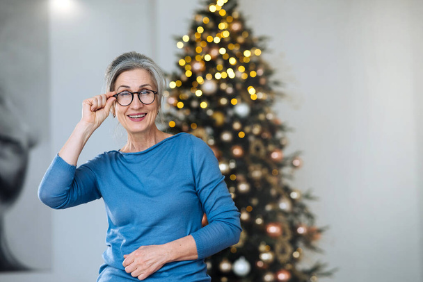 年配の女性の笑顔とカメラを見て、バックグラウンドでクリスマスツリー - 写真・画像