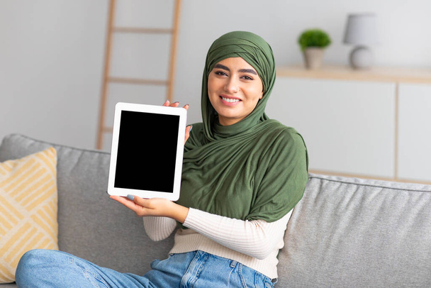 Young Arab lady in hijab showing digital tablet with empty black screen at home, offering space for mockup - Photo, Image