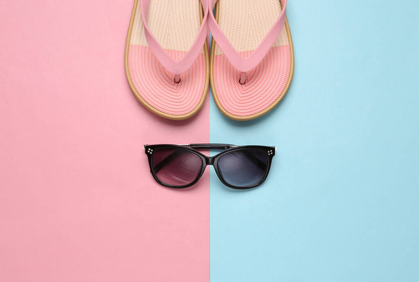 Colored flip flops with sunglasses on blue-pink pastel background. Beach vacation concept. Flat lay - Photo, Image