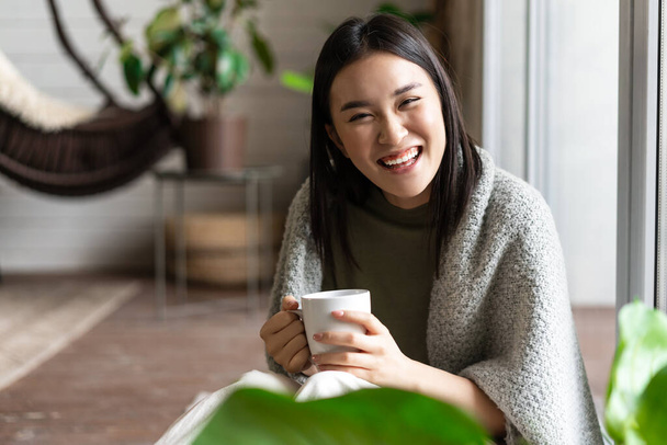 Heureuse fille asiatique riant et souriant, se détendre à la maison, se reposer et boire du thé près de la fenêtre, assis sur le sol dans une couverture confortable - Photo, image