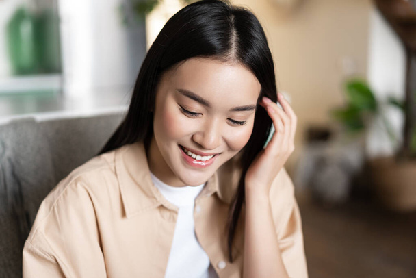 Mignon asiatique fille étudiant souriant, regardant vers le bas et repliant les cheveux derrière l'oreille, assis à la maison à l'intérieur dans des vêtements décontractés - Photo, image