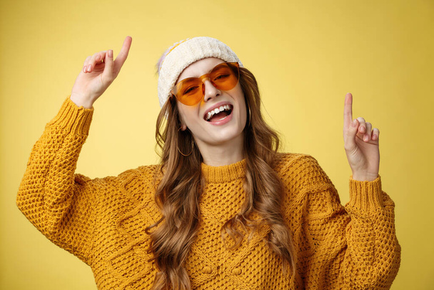 Girl enjoying party have fun smiling broadly happy gazing camera raising index fingers up dancing carefree laughing wearing sunglasses cozy sweater hat, celebrating weekends, yellow background - Photo, Image