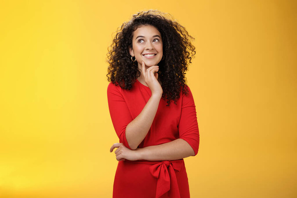 Girl having interesting idea, smiling as feeling confident plan might work. Charming happy woman with curly hair in red dress gazing at upper left corner thoughtful, thinking over yellow wall - Foto, imagen