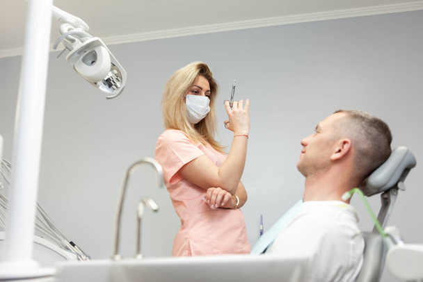 女性歯科医は麻酔注射器を持っています。歯科医院の医務室の椅子に座るハンサムな白人男性 - 写真・画像