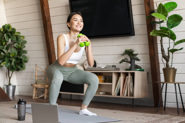 Jovem mulher asiática fazendo exercício em casa com halteres, fazendo agachamentos no tapete de chão, siga on-line guia instrutor de fitness no laptop - Foto, Imagem