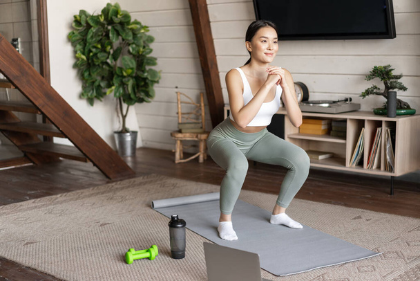 Jovem asiático fitness menina fazendo agachamentos treino em casa, siga on-line esporte ginásio instrutor, de pé no tapete de chão e sorrindo, exercitando-se na sala de estar - Foto, Imagem