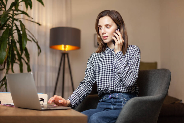 Молода приваблива жінка використовує ноутбук і розмовляє по телефону в її домашньому офісі
 - Фото, зображення