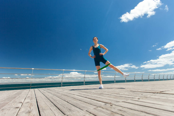Fit γυναίκα κάνει άσκηση με λαστιχάκια γυμναστικής, πόδια κατάρτισης στο ανάχωμα σε φωτεινή ηλιόλουστη μέρα με μπλε ουρανό και σύννεφα. Ευρεία γωνία - Φωτογραφία, εικόνα