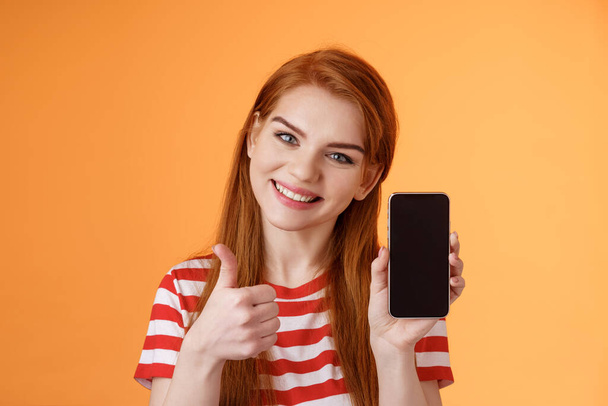 最高のスマートフォンアプリの保証。親切でフレンドリーな赤頭の女性は、携帯電話のアプリケーションをお勧めします親指アップの承認のようなオンラインストアのウェブサイトを紹介し、オレンジの背景に立つ - 写真・画像