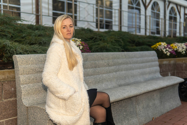portrait of a blonde girl on a bench in a white fur coat in a city park in autumn. - Foto, imagen