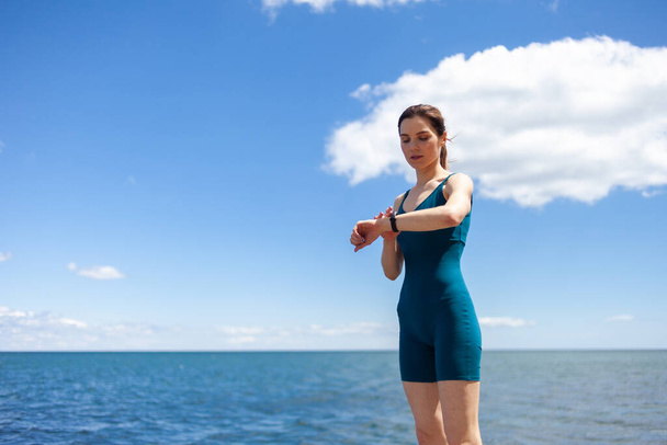 Young fit woman in sports clothes looks at a smart bracelet on the beach against a background of blue sky. Healthy lifestyle - Foto, Imagen