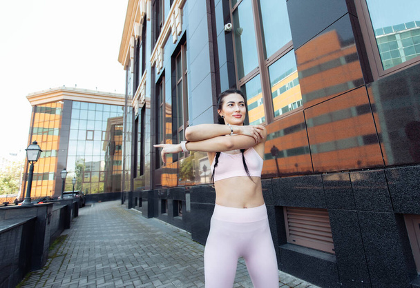 Młoda piękna brunetka z warkoczami ćwiczy rozgrzewkę, rozciągając się przed treningiem na tle budynku miejskiego. Zdrowy styl życia  - Zdjęcie, obraz