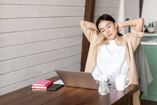 Jeune femme asiatique prend une pause du travail, étirant les bras fatigué de travailler sur un ordinateur portable, termine ses devoirs, étudie à distance de la maison dans la cuisine - Photo, image