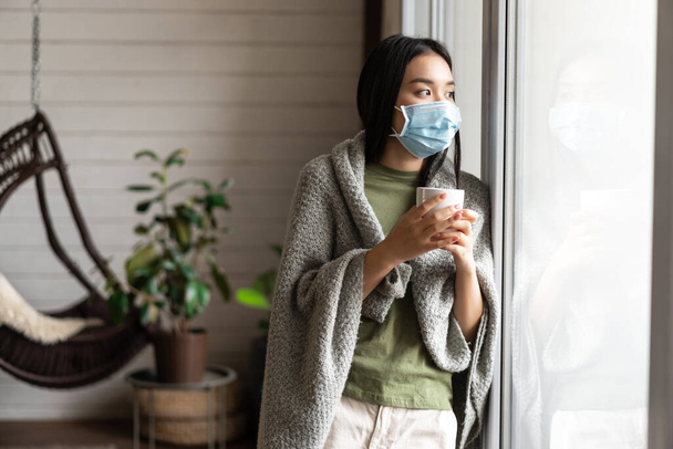 Asian girl in medical face mask, yearning go outside, being sick with covid 19 and staying on self quarantine in her house - Valokuva, kuva
