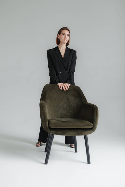 Modeaufnahme einer jungen stilvollen Frau im schwarzen Anzug mit Sessel im Atelier. Vogue, Lifestyle - Foto, Bild