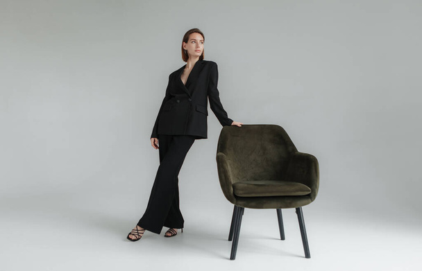 Foto de moda de mujer joven y elegante en traje negro con sillón en el estudio. Vogue, Estilo de vida - Foto, imagen