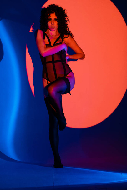 Sensual woman in black underwear with curly hairstyle in studio with neon lights. Sexy portrait of a woman with wavy hair, tan skin and black, transparent bodysuit lingerie in studio. - Foto, Imagem