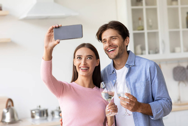 Lächelnd glücklich liebendes junges Paar mit Weingläsern macht Selfie oder Videoanruf in der Küche - Foto, Bild