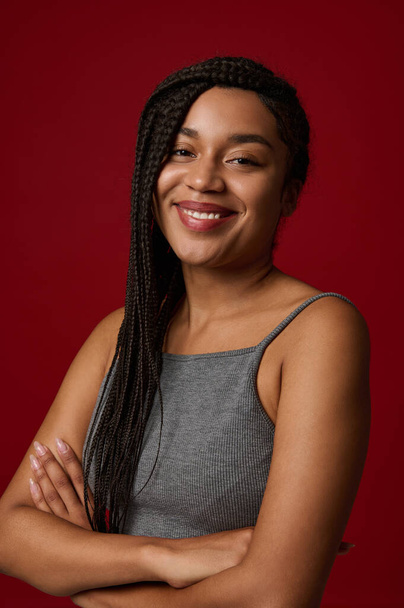 Young adorable African american woman with long pigtails dreadlocks in gray sports t-shirt smiling cute toothy smile while looking at camera, posing against red colored background with copy ad space - Photo, Image