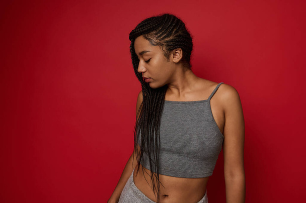 Side portrait of a confident African woman with fit body in gray sportswear with stylish dreadlocks posing looking down on red background with copy space - Foto, immagini