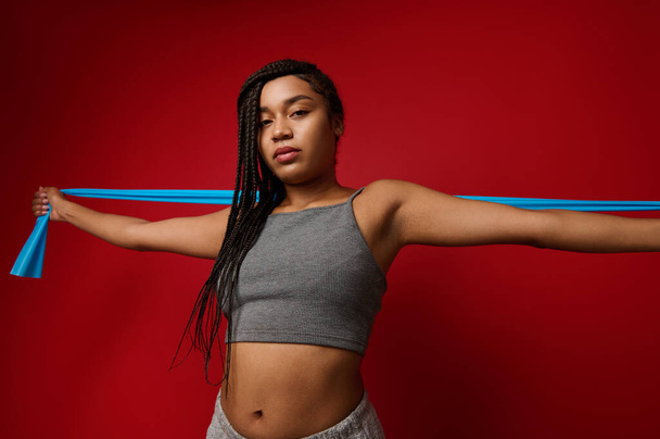 Attractive young African sportswoman wearing wireless headphones, stretching elastic band during her exercise routine, working out against red colored background with space for ad - Photo, Image