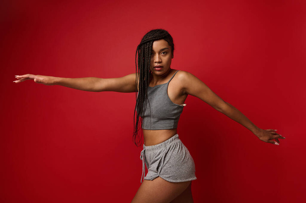 African American sportswoman, determined female athlete with stylish dreadlocks stretches her hands, taking them to the sides, looks at the camera, stretching out on a red background with copy space - 写真・画像