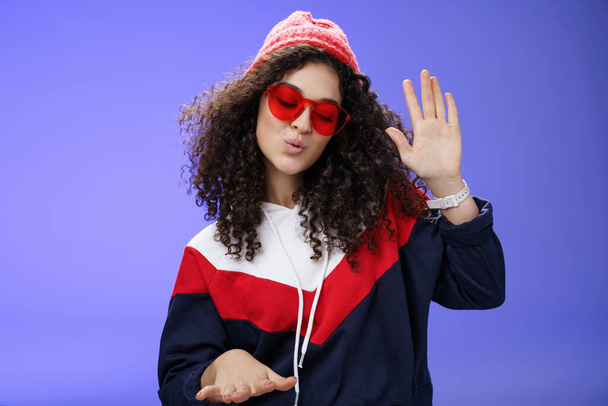 Studio shot of cool and stylish dj girl in red beanie and sunglasses raising palm as enjoying cool music and dancing to rhythm folding lips having fun at awesome party posing chill over blue wall - Photo, Image
