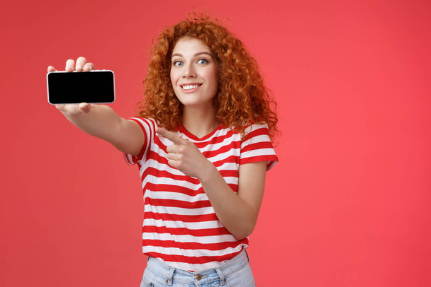 Возбужденный счастливый симпатичный рыжий кудрявый женский шоу горизонтальный экран смартфона указывая дисплей гаджет улыбаясь довольный гордый бить друзей счет игры стоя красный фон - Фото, изображение