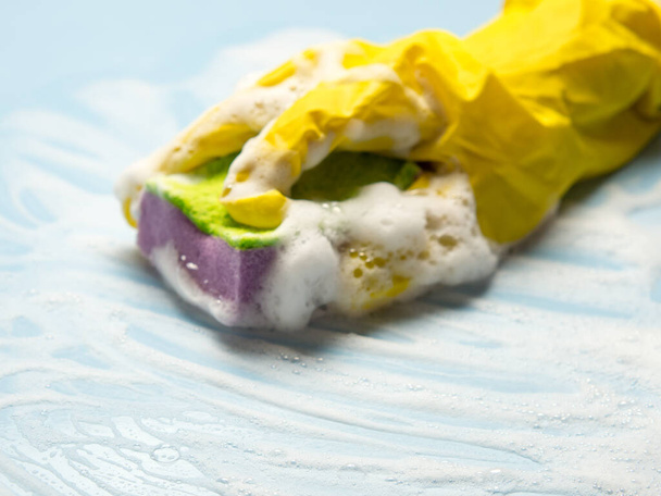 Una mano en un guante de goma amarillo sostiene una esponja de limpieza en espuma jabonosa sobre un fondo azul. concepto de limpieza de la casa, servicio de limpieza - Foto, imagen