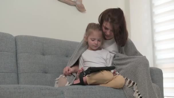 Een moeder met een dochtertje zit op de bank en speelt op de tablet verpakt in een deken - Video