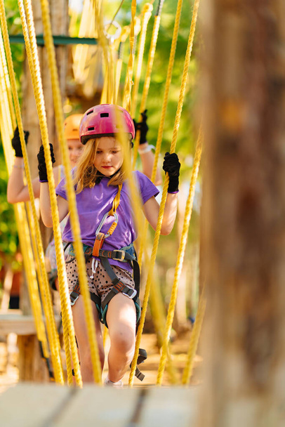 girl in helmet, T-shirt and shorts climbs in rope park - Фото, изображение