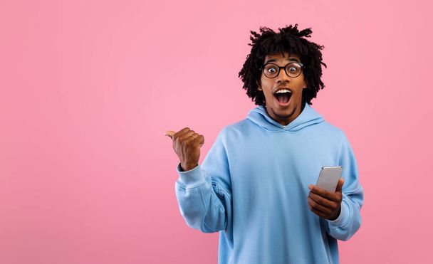 Izgalmas fekete tini srác használ mobiltelefon, reklám hűvös alkalmazás, rámutatva az üres hely rózsaszín háttér - Fotó, kép