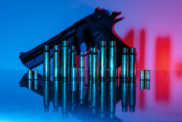 Pistola de mano con una fila de carcasas de municiones de latón en la parte delantera, colorido iluminado en azul y rojo y un reflejo simétrico espejo - Foto, imagen