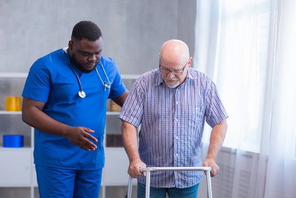 Le soignant apprend au vieil homme à marcher avec un marcheur. Infirmière professionnelle et patiente dans une maison de retraite. Assistance, réhabilitation et santé. - Photo, image