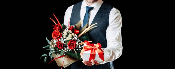 Gift box hand. Happy young business man holding surprise giftbox present with luxury rose flower bouquet isolated on black background. Valentines day, Christmas, New Year banner with copy space - Photo, Image