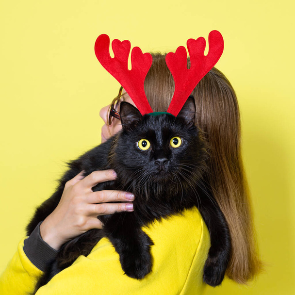 Κορίτσι σε ένα κίτρινο φόρεμα κρατά την αγαπημένη χνουδωτή αστεία μαύρη γάτα της με κίτρινα μάτια σε χριστουγεννιάτικο τάρανδο Antlers Headband σε ένα κίτρινο φόντο. Χριστουγεννιάτικη γάτα. - Φωτογραφία, εικόνα