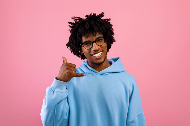 Веселый афро-американский подросток жестом зовет меня, держа за руку, как телефон рядом с ухом на розовом фоне студии - Фото, изображение