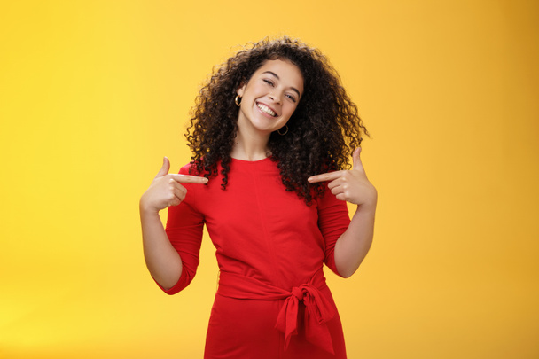 Orgullosa y satisfecha ambiciosa estudiante exitosa en vestido rojo de pie contenta sonriendo y señalándose a sí misma como si presumiera de sus propios logros felizmente y contenta sobre la pared amarilla - Foto, Imagen
