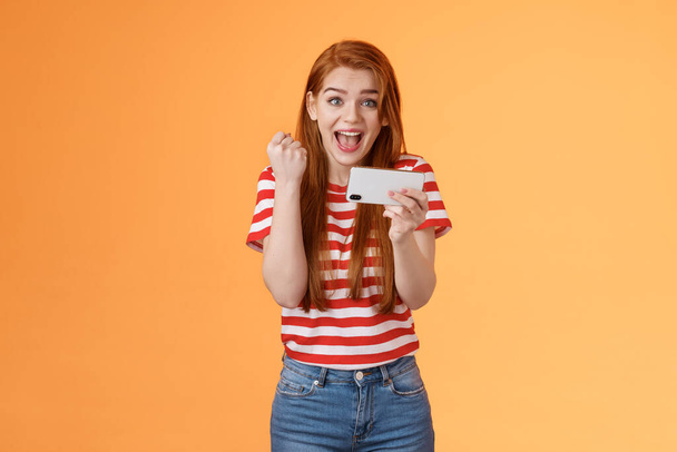 Happy excited redhead female pass level, like awesome game, score goal, hold smartphone horizontally, fist pump success, celebrate lucky achievement, smiling broadly, orange background - Foto, immagini