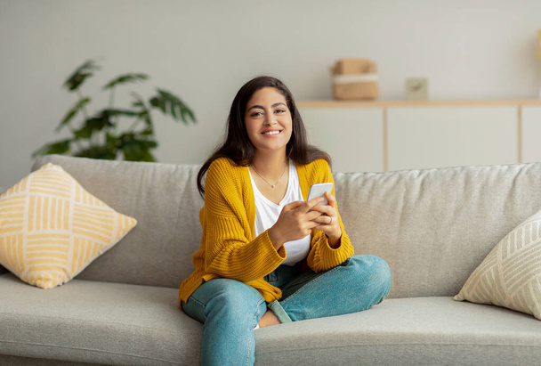 Senhora do Oriente Médio positivo sentado no sofá e usando smartphone, conversando com amigos ou amante, sorrindo para a câmera - Foto, Imagem