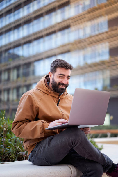 Vertical photo of a man using laptop outdoors. Businessman with a nice smile works remotely on the street. - Photo, Image
