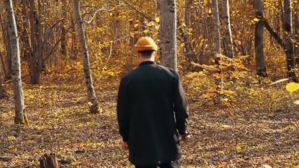一人の若い男は黒いジャケットを着て、黄色の帽子は落ち葉で美しい森の中の木の間を歩く。自然な背景。バックビュー。秋だ。屋外だ。スローモーション - 映像、動画
