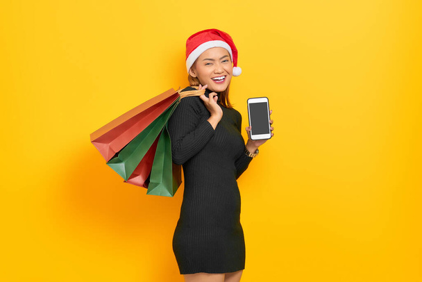Smiling young Asian woman in Santa Claus hat showing blank screen mobile phone and holding shopping bags isolated over yellow background - Valokuva, kuva