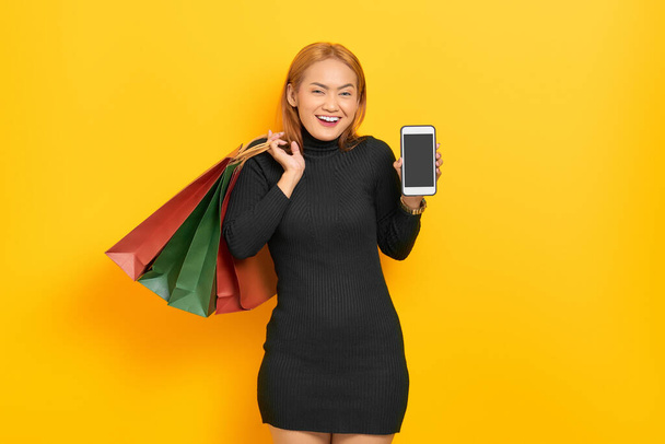 Usmívající se mladá asijská žena ukazující prázdné obrazovce mobilní telefon a držení nákupních tašek izolované přes žluté pozadí - Fotografie, Obrázek