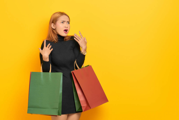 Surprised young Asian woman holding shopping bags and looking away at copy space isolated over yellow background - 写真・画像