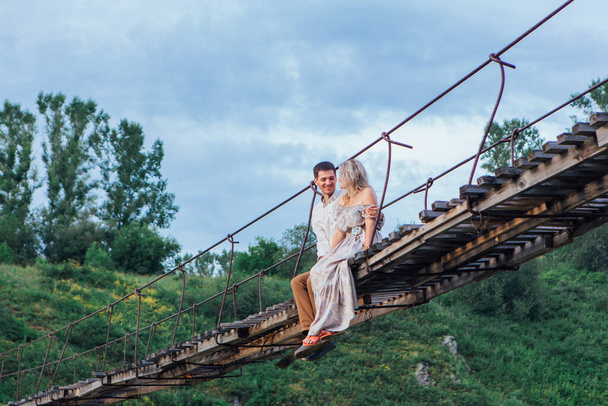 Beautiful young romantic couple sitting and hugging on the suspension bridge above the river - Fotografie, Obrázek