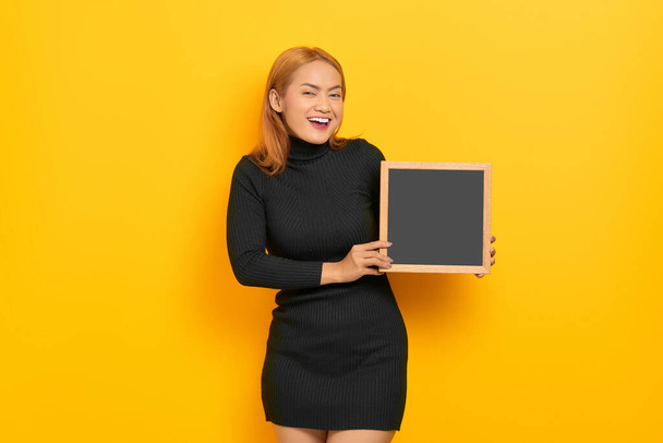 Sorridente giovane donna asiatica che tiene lavagna isolata su sfondo giallo - Foto, immagini