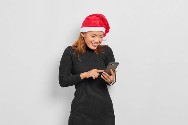 Портрет усмішки молодої азіатки у капелюсі Санта - Клауса за допомогою мобільного телефону, ізольованого на білому тлі. - Фото, зображення